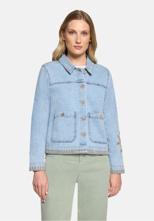 Femme portant une veste en jean bleu clair avec des fermetures à boutons et des coutures décoratives, associée à un pantalon vert clair, face à l'avant.
