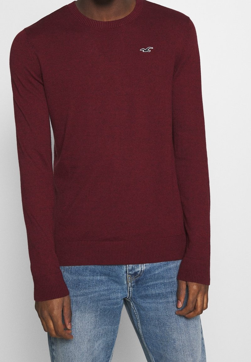 Homme portant un pull bordeaux à manches longues avec un petit logo d'oiseau blanc sur la poitrine et un jean bleu clair.
