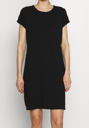 Kobieta ubrana w prostą czarną sukienkę z krótkim rękawem, okrągłym dekoltem i małą kieszonką na piersi, stojąca na jednolitym tle.