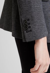Manica di un blazer a motivo houndstooth bianco e nero con tre bottoni neri. Il tessuto sembra testurizzato, aderente lungo il polso.