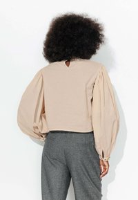 Blouse beige cropped avec des manches bouffantes, présentant un détail de dos en forme de trou de serrure. Texture lisse, associée à un pantalon à carreaux gris.