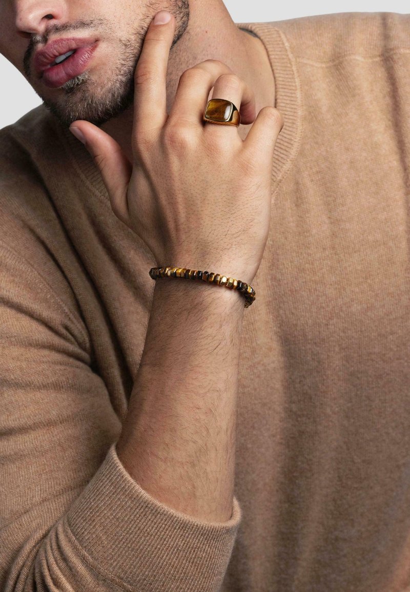 Brun strikket sweater, hånd på hagen, bærer en guldring med en stor sten og et perlesmykke med sekskantede former i sort og guld.