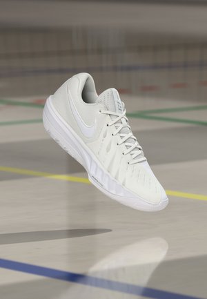 Λευκό αθλητικό παπούτσι Nike με επάνω μέρος από διχτυωτό υλικό και κορδόνια, αιωρούμενο στον αέρα πάνω από το δάπεδο ενός γυμναστηρίου με έγχρωμες γραμμές γηπέδου.