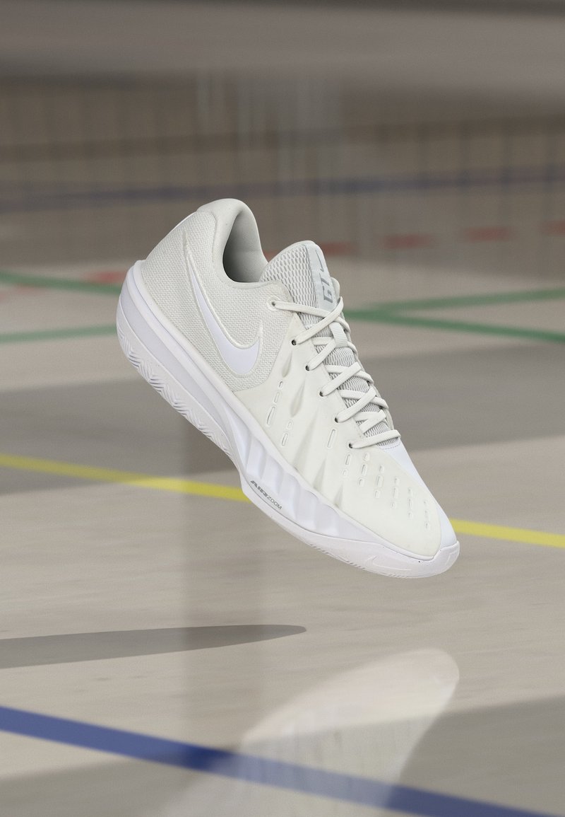 Λευκό αθλητικό παπούτσι Nike με επάνω μέρος από διχτυωτό υλικό και κορδόνια, αιωρούμενο στον αέρα πάνω από το δάπεδο ενός γυμναστηρίου με έγχρωμες γραμμές γηπέδου.