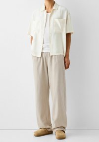 Korte mouw crème shirt, twee zijzakken, witte onderhemd, licht beige broek en tan instappers. Soepele stof, comfortabele fit.