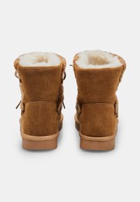 Stivaletti alla caviglia in suede marrone con una morbida fodera in pelliccia color crema e lacci decorativi laterali. Suole in gomma resistenti offrono aderenza e comfort.