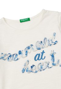 Camisa de algodón blanca con texto azul que dice "mould at heart", con un diseño texturizado y ondulado, y un acabado suave y liso. Etiqueta de la marca en verde visible.