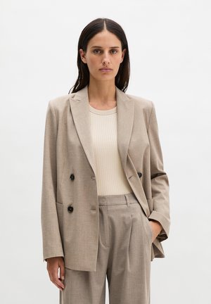 Frau steht mit der Hand in der Tasche, trägt beige zweireihigen Blazer, passende Hose und cremefarbenes geripptes Oberteil vor weißem Hintergrund.