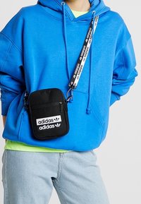 Černá kabelka přes rameno se dvěma bílými logy adidas, s uzavíratelnou přihrádkou a vzorovaným nastavitelným popruhem, nošena přes modrou mikinu s kapucí.