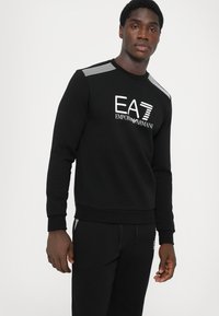 Black sweatshirt featuring a white EA7 Emporio Armani logo. Design includes striped accents on shoulders and ribbed cuffs. Cotton blend fabric.