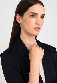 Sudraba aproce ar smalku ķēdes dizainu, kurā ir mazi caurspīdīgi akmeņi, uzvilkta uz rokas. Modelis valkā tumšu žaketi un gaišu džemperi.