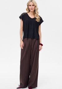 Czarny top z krótkim rękawem i dekoltem w kształcie litery V, ozdobiony detalami na ramionach, zestawiony z szerokimi brązowymi spodniami i bordowymi butami w szpic.