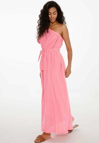 Ružová maxi šaty na jedno plece vyrobené z ľahkého materiálu, s viazaným pásom, voľnou siluetou a jemnou textúrou. Nosené hnedé sandále.