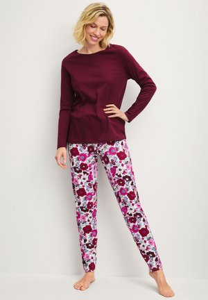 Frau trägt ein langärmeliges dunkelrotes Oberteil und weiße Leggings mit pinken und burgunderfarbenen Blumenmustern, steht barfuß an einer weißen Wand.