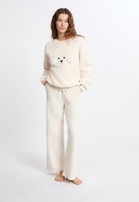 Felpa di colore crema con un volto di orso testurizzato e orecchie. Abbinata a pantaloni larghi color avorio. Design morbido e rilassato.