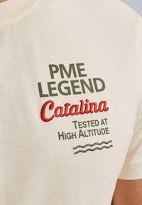 T-shirt en coton beige avec texte brodé vert et rouge. Le texte indique "PME LEGEND", "Catalina" et "Testé en haute altitude". Détail de ligne ondulée.