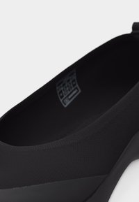 Μαύρο παπούτσι χωρίς κορδόνια με υφασμάτινο επάνω μέρος με υφή, στρογγυλεμένη μυτερή μύτη και χαμηλό τακούνι, εμφανίζεται σε κοντινή λήψη από λοξή επάνω οπτική γωνία.