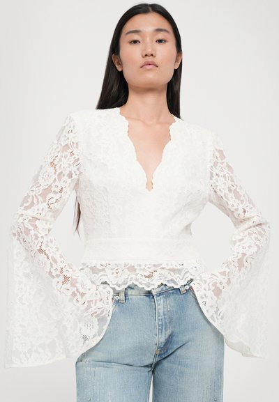 Witte kanten top met een diepe V-hals en bell mouwen, versierd met bloemenpatronen en een scalloped zoom, gecombineerd met lichtblauwe denim jeans.