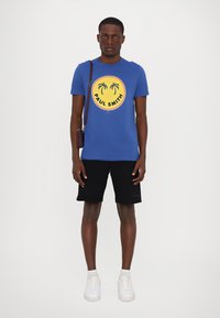 Zila kokvilnas T-krekls ar apaļu dzeltenu logotipu, kurā attēlotas palmas un "Paul Smith". Apģērbts ar melniem šortiem un baltiem sporta apaviem.