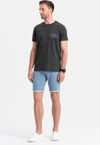 Camiseta negra de manga corta con un bolsillo en el pecho, combinada con pantalones cortos de mezclilla azul claro doblados en el dobladillo. Zapatillas blancas tipo slip-on.