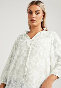 Top bianco e trasparente con ricami floreali, maniche lunghe e scollatura con laccetti. Tessuto morbido con una vestibilità rilassata e dettagli arricciati.
