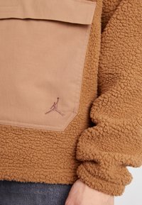 Casaco de fleece castanho com uma superfície texturizada, apresentando um bolso bege e um logotipo bordado. As mangas são largas e arredondadas, aumentando o conforto.