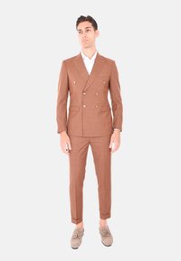 Completo marrone doppiopetto con spalle strutturate, due tasche frontali, pantaloni slim fit, abbinato a mocassini beige e una camicia bianca.