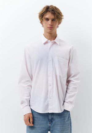 Mladý muž s rozcuchanými blond vlasmi, oblečený v voľnej bielej košeli na gombíky a modrých džínsach, stojaci pred jednoduchým bielym pozadím.