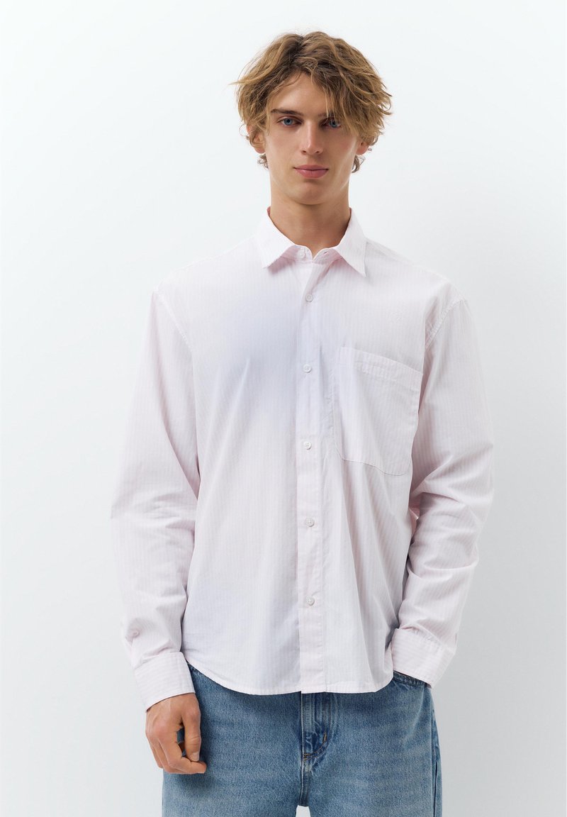 Jeune homme aux cheveux blonds ébouriffés portant une chemise blanche ample à boutons et un jean bleu, debout devant un fond blanc uni.
