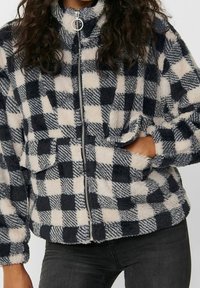 Veste en polaire à motif à carreaux bleu marine et crème, fermeture éclair, col montant et poches avant. Texture douce avec des nuances variées.