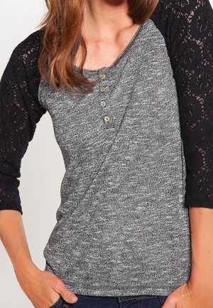 Femme portant un haut en maille grise avec des manches trois-quarts en dentelle noire et quatre petits boutons près de l'encolure.