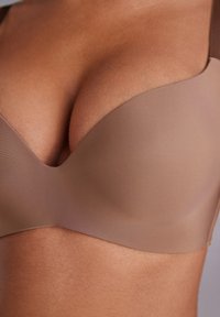 Bruine gewatteerde beha met gladde stof, naadloze cups en een brede onderband. Het heeft een laag uitgesneden, gevormd ontwerp voor een verbeterde vorm.