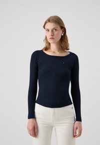 Marineblaues, geripptes Langarmoberteil mit Rundhalsausschnitt, versehen mit einem dezenten Logoakzent auf der Brust. Kombiniert mit weißen Hosen.