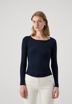 Maglione a maniche lunghe blu navy a costine con scollo rotondo, caratterizzato da un sottile logo sul petto. Abbinato a pantaloni bianchi.