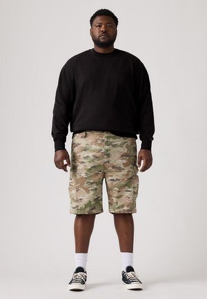 Un hombre está de pie mirando al frente, con una sudadera negra, pantalones cortos cargo de camuflaje, calcetines blancos y zapatillas negras, sobre un fondo liso y claro.