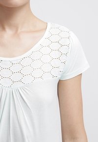 Camisa de manga corta de color azul claro con detalles de encaje en la parte delantera, que presenta un patrón ovalado y tejido fruncido en la parte inferior. Textura suave.