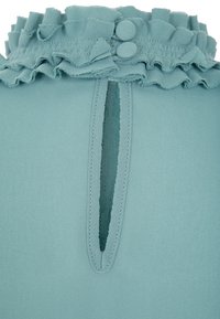 Blusa teal con scollo a volant, dotata di due bottoni rivestiti in tessuto. Dettaglio dello spacco posteriore che corre verticalmente sotto il bordo a volant.