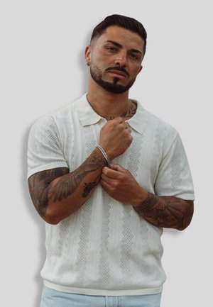 White short-sleeve polo shirt with textured zigzag pattern, featuring a collar and ribbed cuffs. Light blue jeans and visible tattoos on arms.