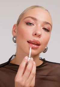 Femme aux cheveux blonds tirés en arrière et boucles d'oreilles métalliques, appliquant un gloss rose brillant, vêtue d'un haut noir transparent devant un fond uni.