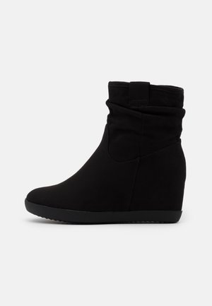 Bottine noire à cheville avec talon compensé, dessus souple et ample, et détail d'onglet sur un fond clair uni.