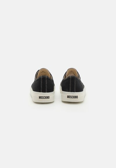 Svarta canvas slip-on skor med vit söm och gummisula, med en logotagslabel på hälen. Slät textur och rundad design.