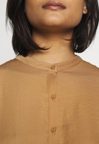Brown button-up blouse with a soft, smooth texture, featuring a round neckline and three brown buttons down the front.