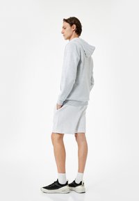 Conjunto de sudadera gris claro y pantalones cortos. La sudadera tiene capucha con cordones; el modelo lleva zapatillas negras con suelas blancas y calcetines de longitud media.