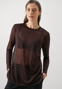 Hosszú ujjú, félig áttetsző barna felső kerek nyakkivágással. Simább textúra és ívelt szegély, fekete melltartóra rétegezve. Egyszerű dizájn.