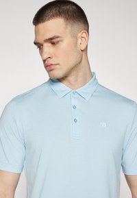 Lys blå polo skjorte med en myk tekstur, tre-knappers stolpe og en liten brodert logo på venstre bryst. Korte ermer.