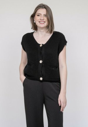 Femme souriante, portant un cardigan noir en maille sans manches avec de gros boutons et des poches, et un pantalon noir, debout devant un fond clair.