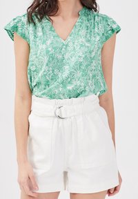 Blouse à imprimé floral vert avec un col en V et des manches volantées, associée à un short blanc taille haute avec une ceinture et des détails en métal argenté.