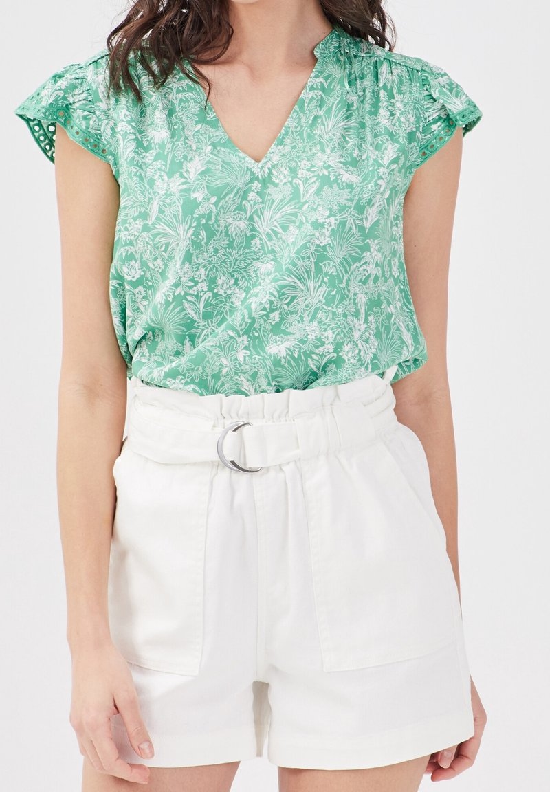 Blouse à imprimé floral vert avec un col en V et des manches volantées, associée à un short blanc taille haute avec une ceinture et des détails en métal argenté.