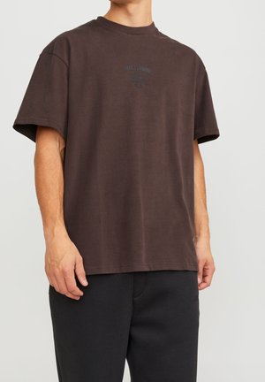 Mand iført løs mørkebrun T-shirt med lille tekstlogo på brystet og sorte bukser, stående mod en ensartet lys baggrund.