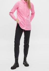 Woman stands wearing a bright pink button-up shirt, black slim-fit jeans, and black ankle boots on a plain white background.